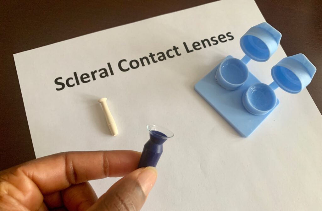 A contact lens case sits on a paper labeled "Scleral Contact Lenses," as a hand in the foreground grips a scleral lens suction tool, carrying a scleral lens.