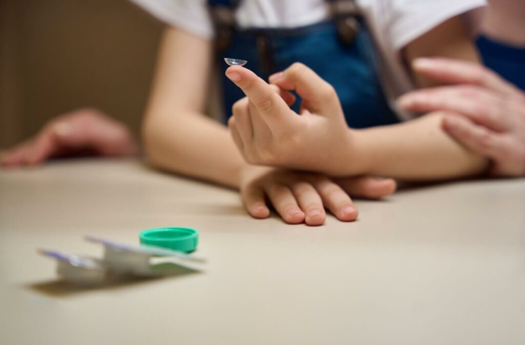 Below-the-face shot of a child holding a contact lens on the tip of their finger with a parent's hand on their arm. Contact lens case in the foreground.