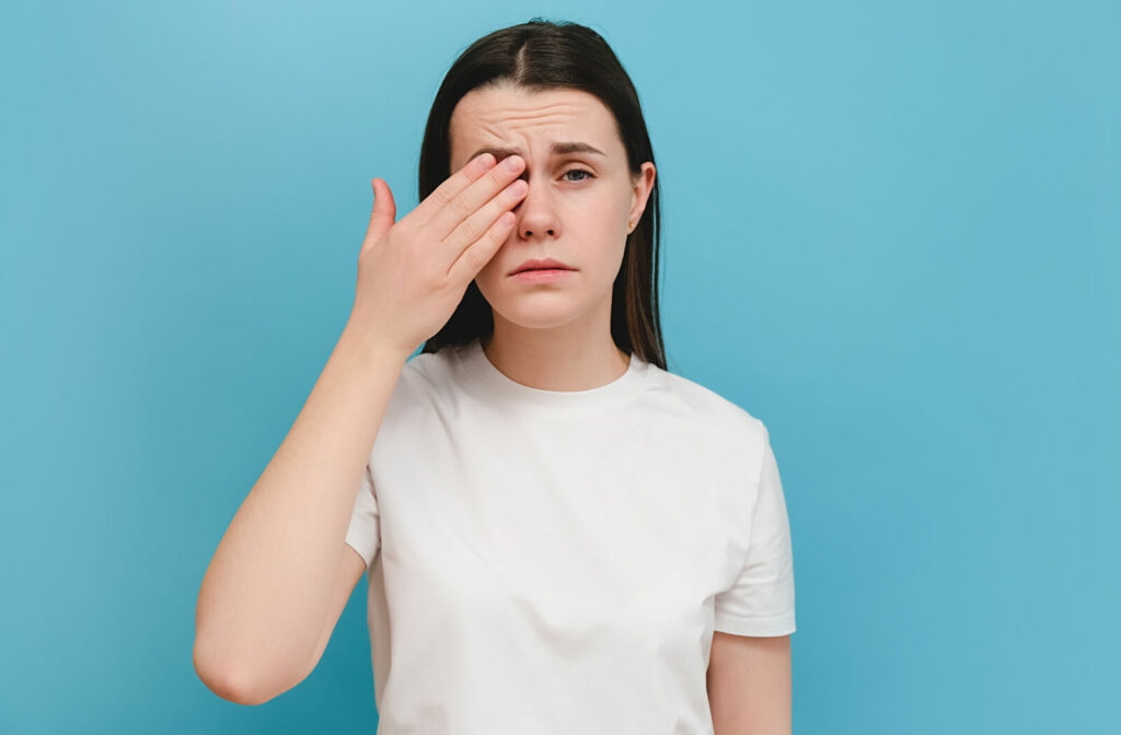 A person in a white t-shirt stands against a blue background, rubbing her eye with a pained expression, showing eye discomfort.