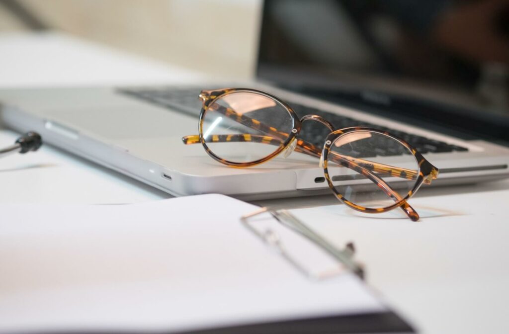 Pair of eyeglasses resting on a laptop computer next to paperwork on a desk.