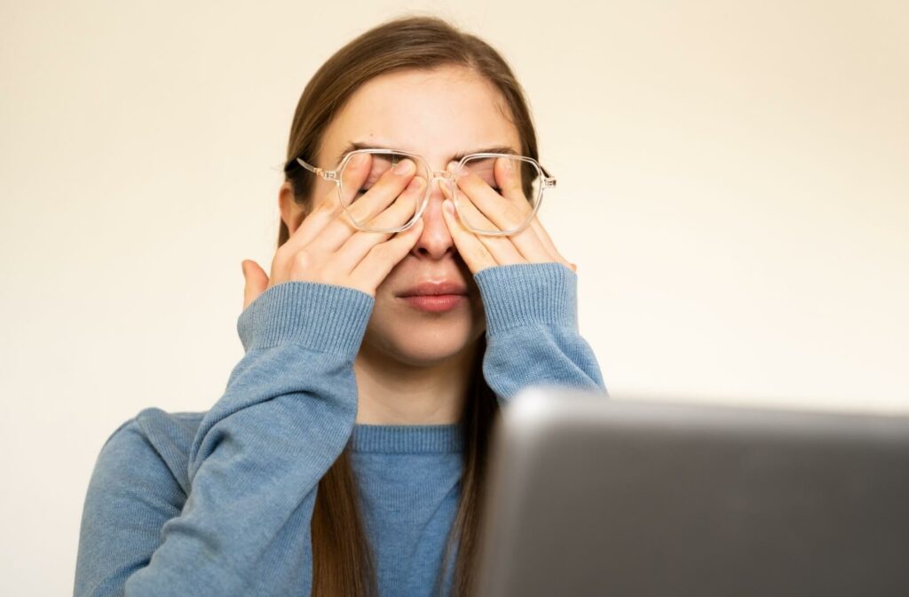 A person rubbing the eyes in front of a laptop while wearing eyeglasses, illustrating digital eye strain and blurred vision from dry eyes.