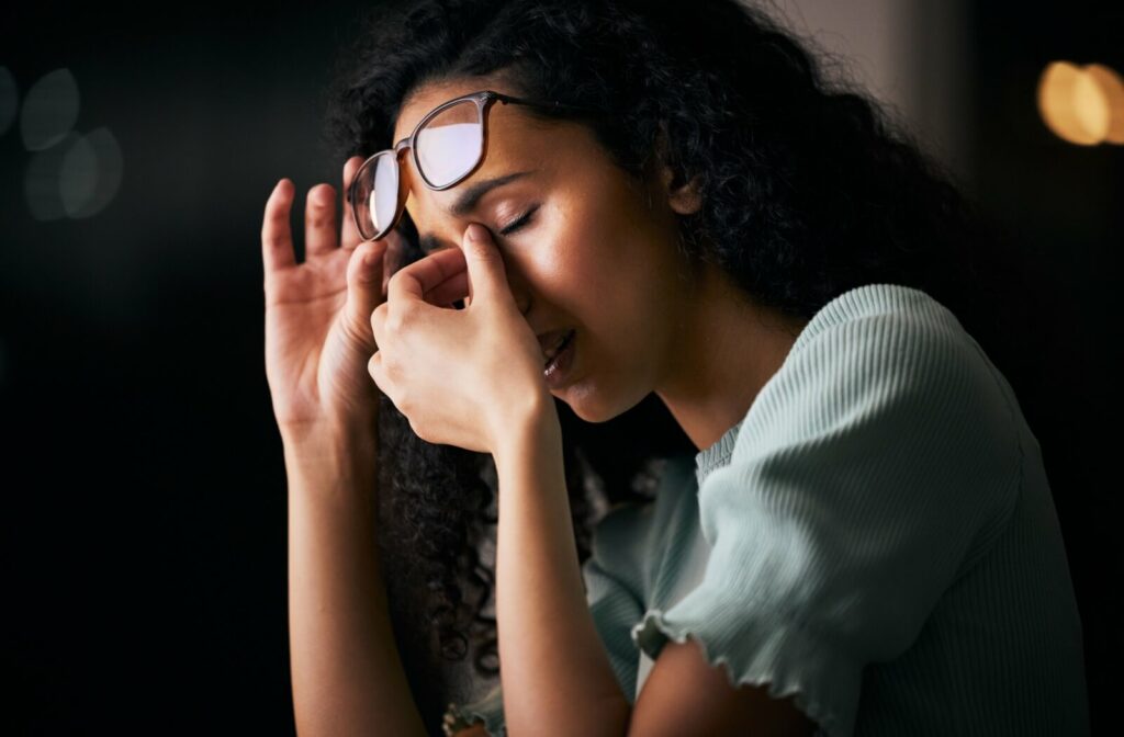 A young woman with curly hair lifting her eyeglasses to rub the bridge of her nose and closed eyes in a dimly lit room.