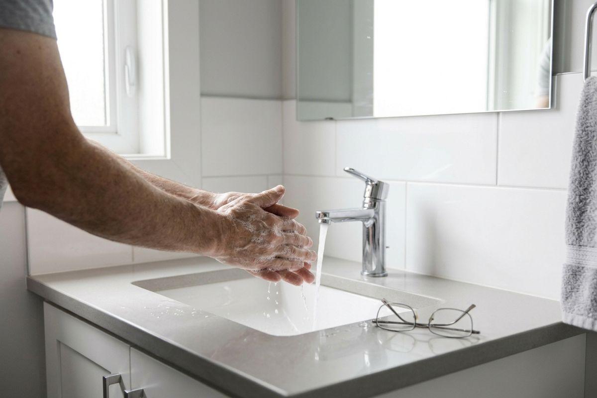 Person washing soapy hands under running water at a white bathroom sink, with glasses resting on the countertop and a towel nearby.